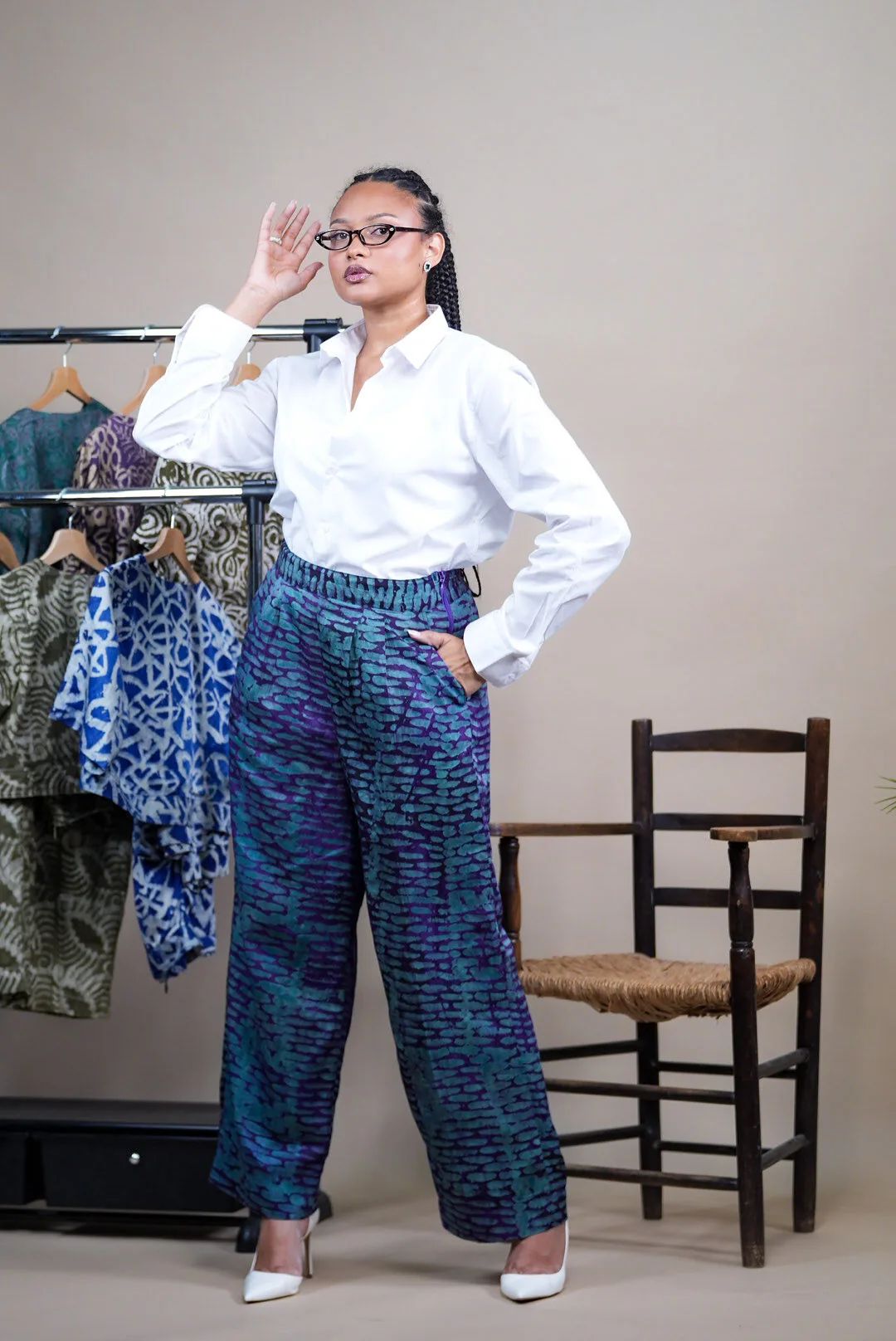 Femme à lunettes en chemise blanche et pantalon Adire bleu violet, devant un portant